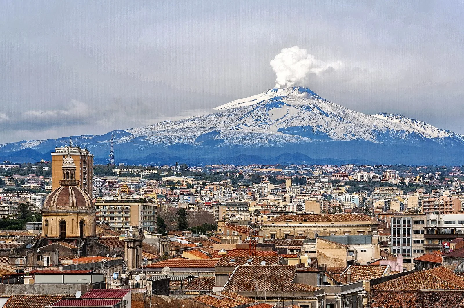 Il centro storico di Catania visto dall'alto, con cupole e tetti in primo piano e l'Etna innevata sullo sfondo