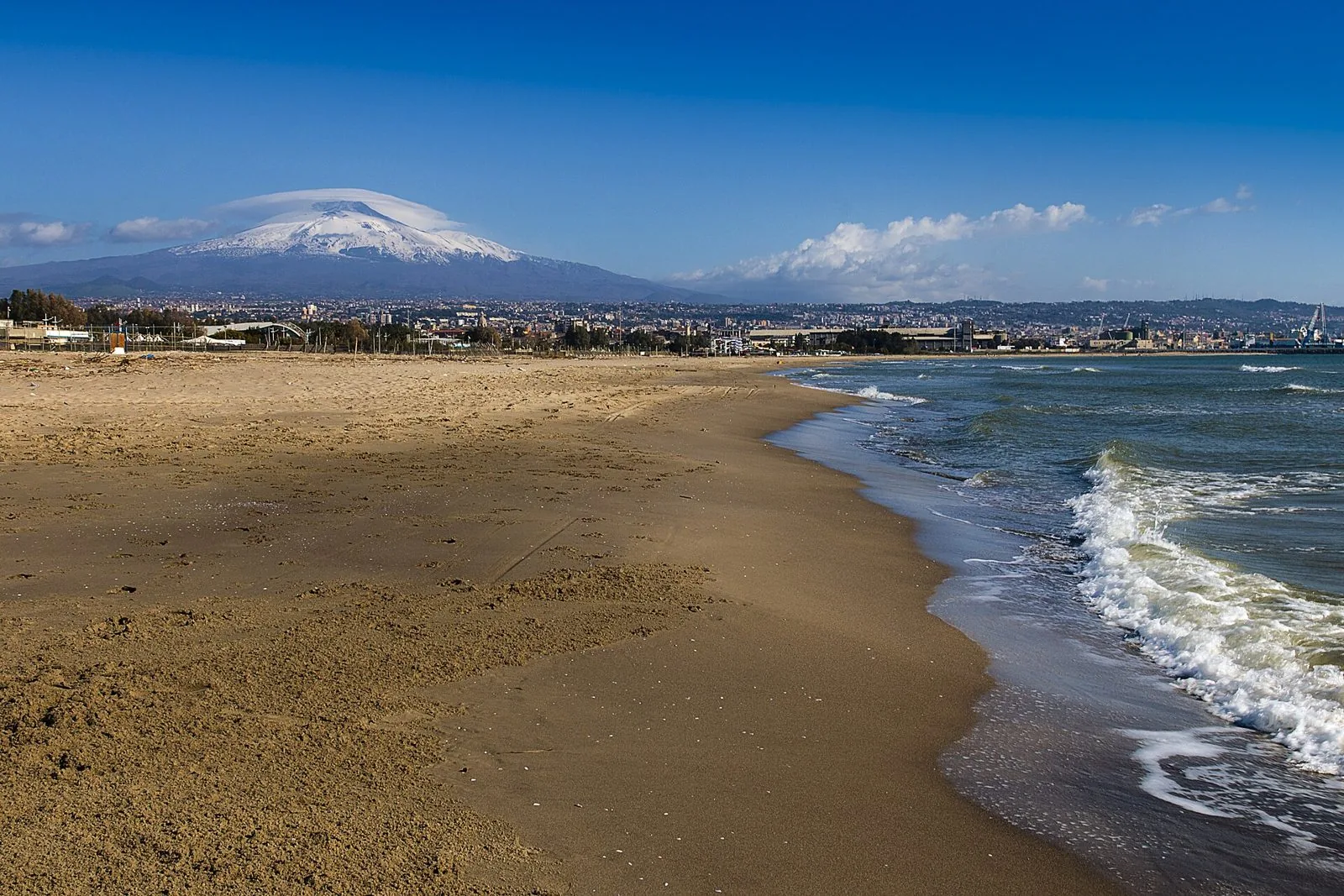 La spiaggia sabbiosa della Playa di Catania con le onde sulla riva e, sullo sfondo, la città e l'Etna innevato sotto un cielo blu