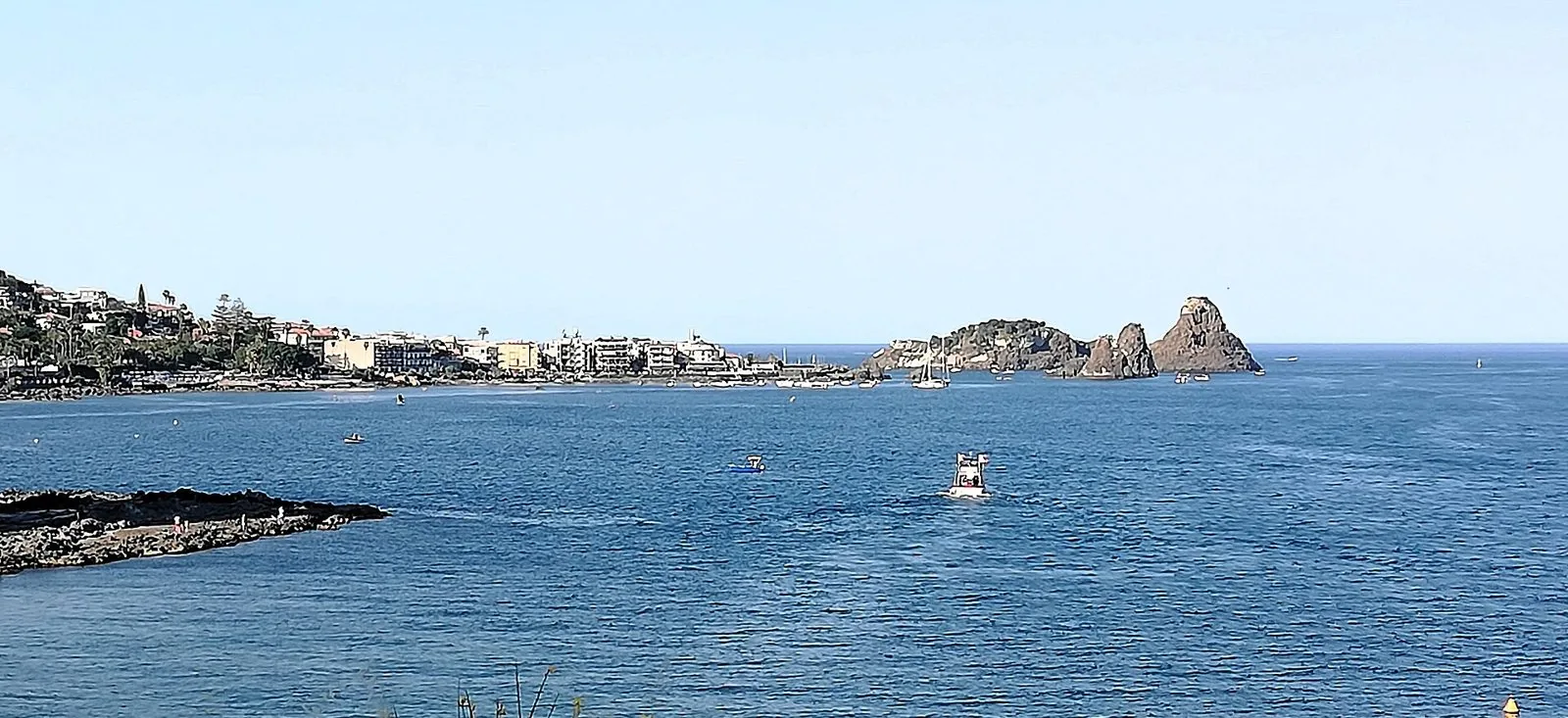 I Faraglioni dei Ciclopi davanti ad Aci Trezza visti dalla costa di Aci Castello, con il paese affacciato sul mare e alcune barche nell'acqua blu